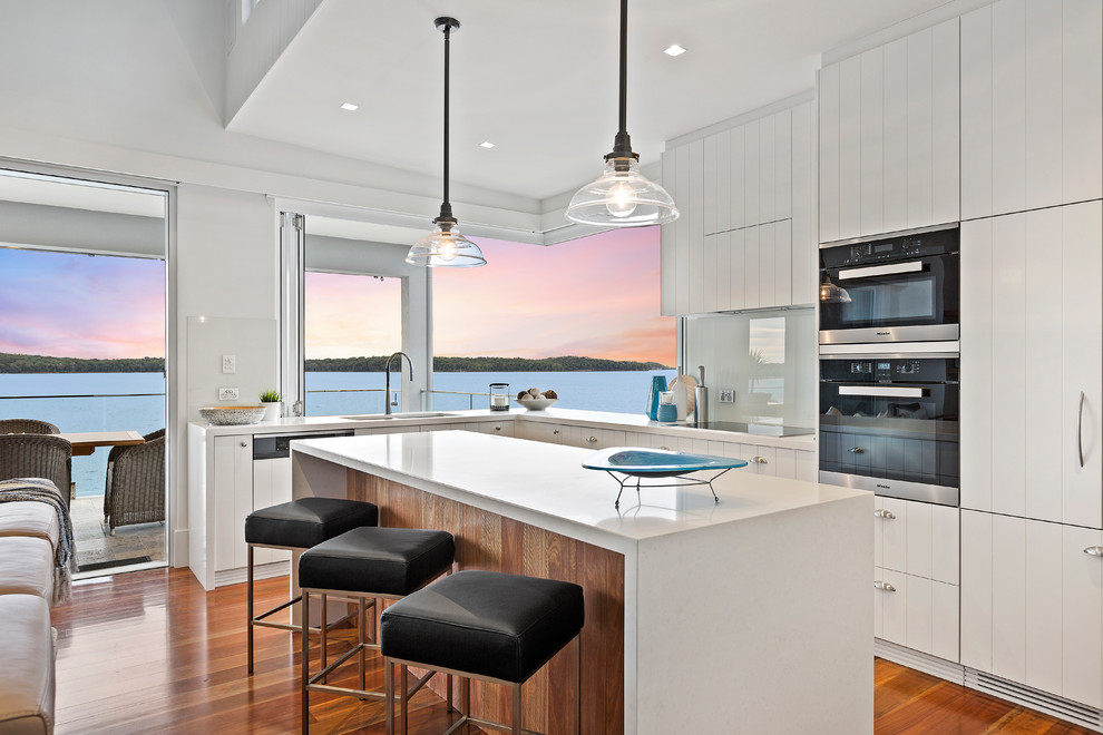 Photo of a contemporary l-shaped open plan kitchen in Central Coast with quartz benchtops, a drop-in sink, flat-panel cabinets, white cabinets, glass sheet splashback, black appliances, medium hardwood floors and with island.