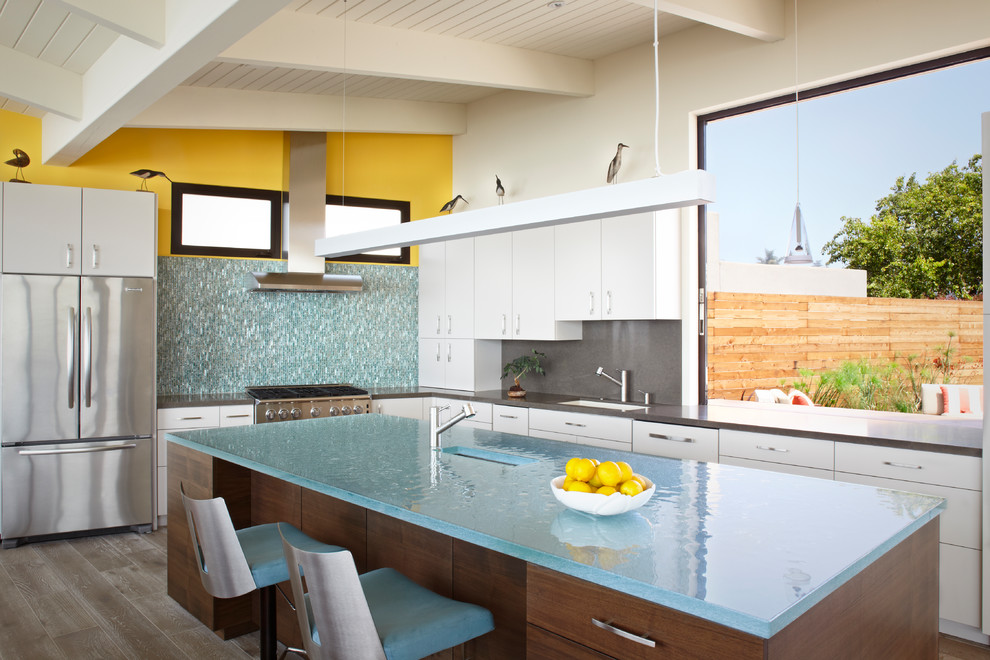 Inspiration for a mid-sized midcentury l-shaped kitchen in Santa Barbara with an undermount sink, flat-panel cabinets, white cabinets, glass benchtops, stainless steel appliances, with island, grey splashback, dark hardwood floors, brown floor and blue benchtop.