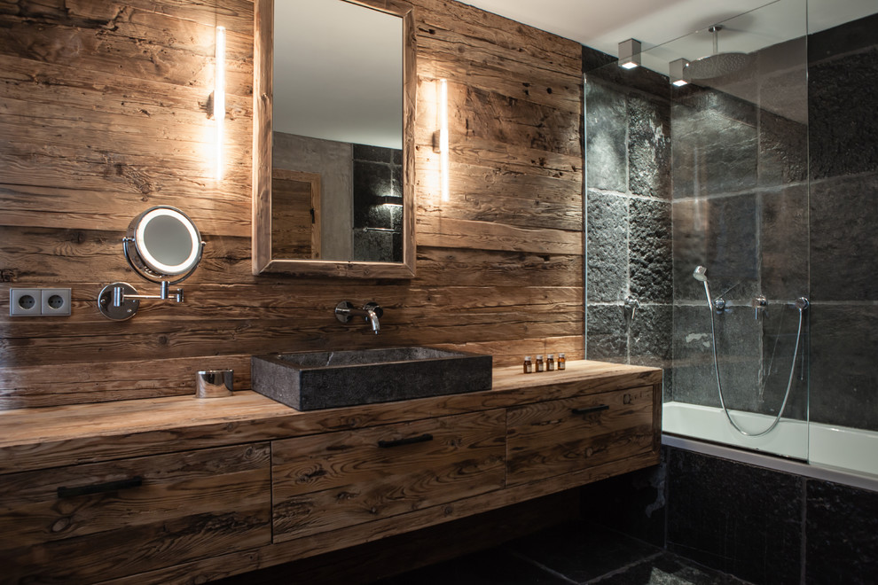 Inspiration for a mid-sized country 3/4 bathroom in Munich with flat-panel cabinets, medium wood cabinets, a drop-in tub, a shower/bathtub combo, black tile, slate, brown walls, slate floors, a vessel sink, wood benchtops, black floor and a hinged shower door.