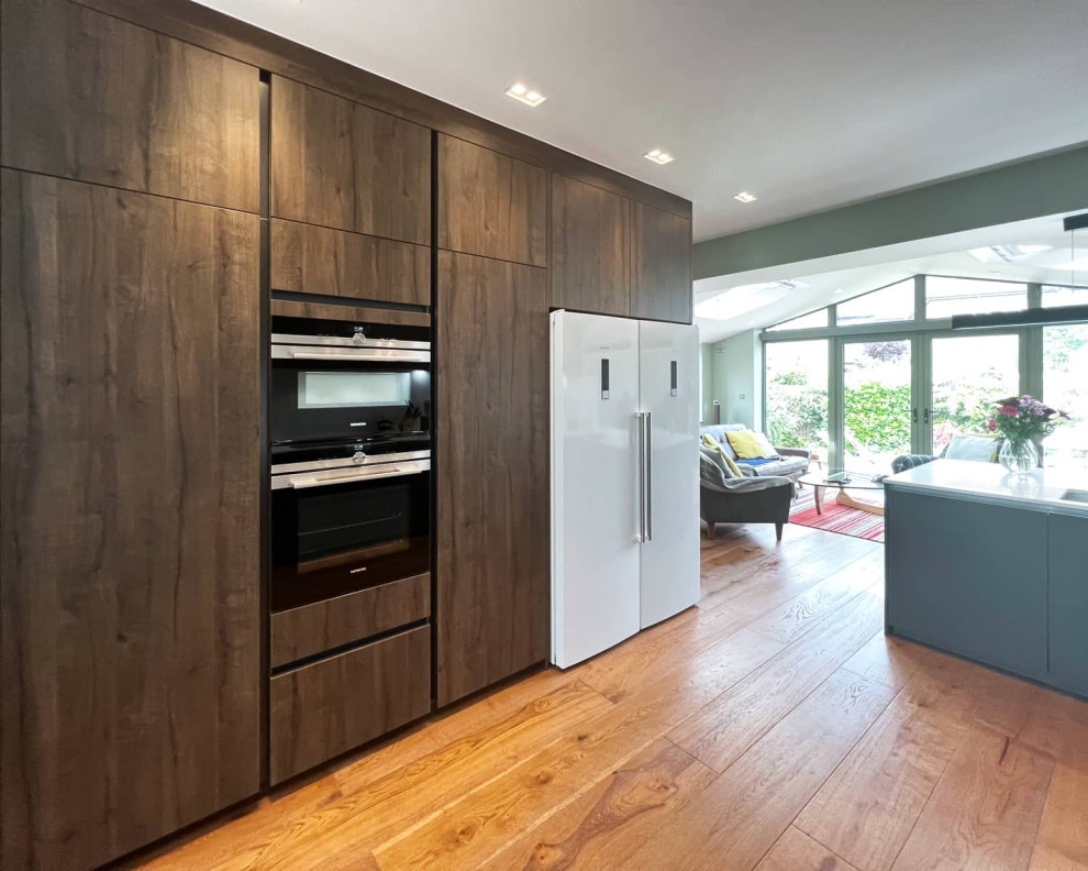 Mid-sized contemporary galley eat-in kitchen in London with a single-bowl sink, flat-panel cabinets, grey cabinets, quartzite benchtops, white splashback, black appliances, light hardwood floors, brown floor and white benchtop.