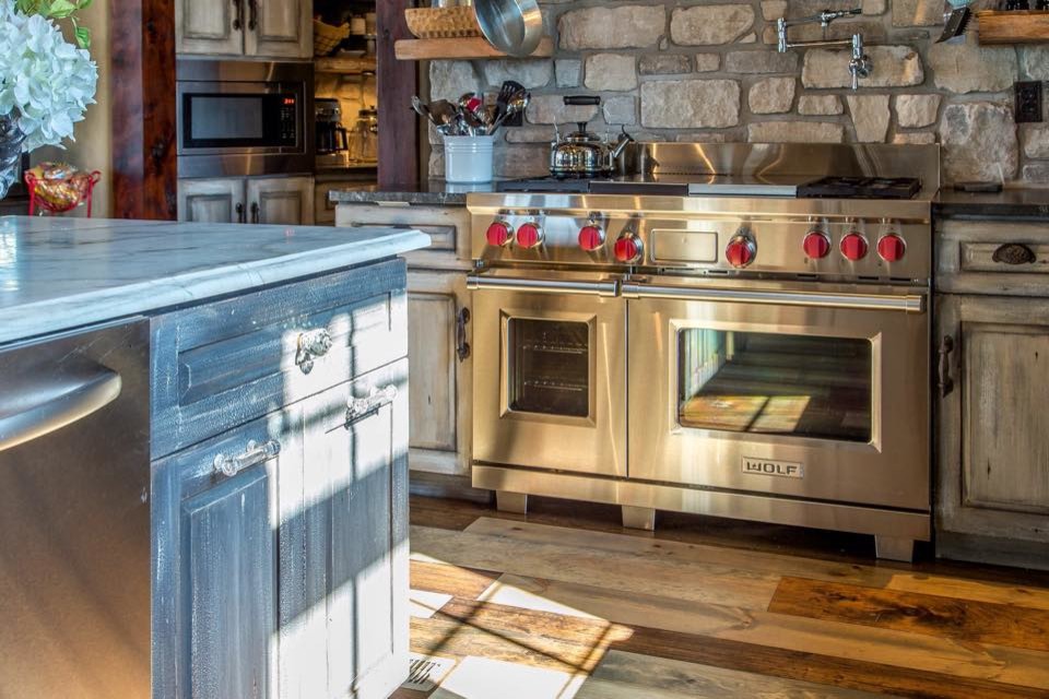 Eat-in kitchen - mid-sized rustic l-shaped medium tone wood floor and brown floor eat-in kitchen idea in St Louis with a farmhouse sink, distressed cabinets, gray backsplash, stainless steel appliances, an island, raised-panel cabinets, marble countertops and stone tile backsplash