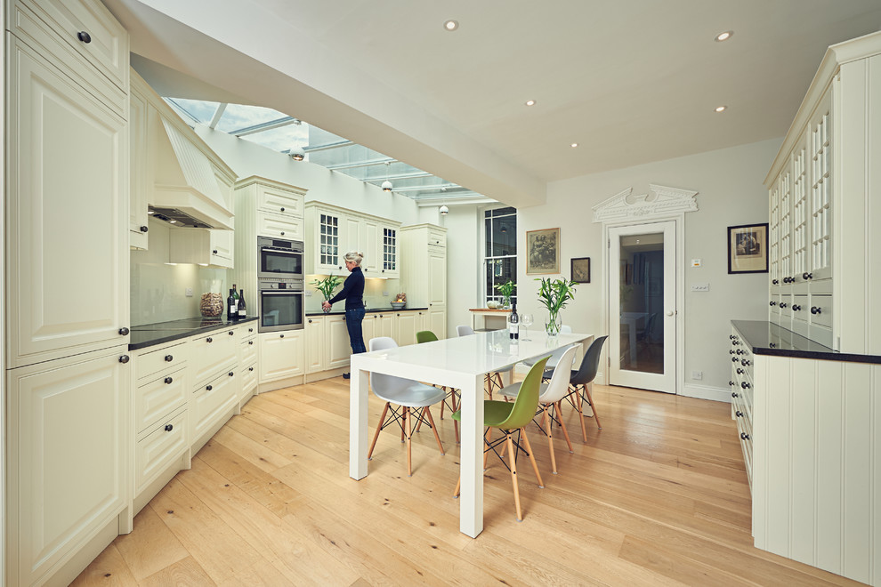 Large eclectic u-shaped eat-in kitchen in London with an integrated sink, raised-panel cabinets, white cabinets, granite benchtops, white splashback, glass sheet splashback, black appliances and light hardwood floors.