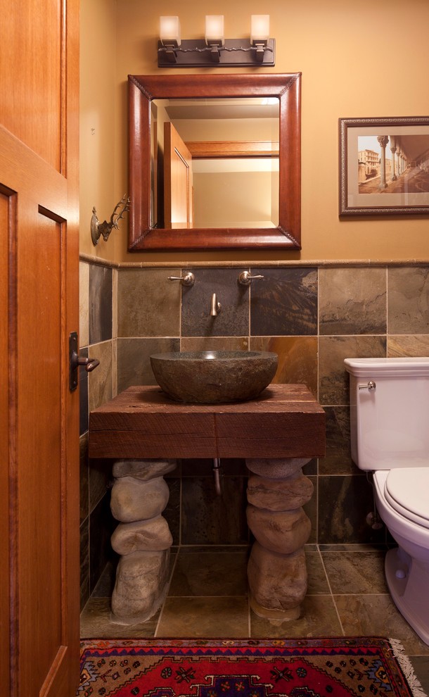 Mid-sized traditional powder room in Minneapolis with a vessel sink, wood benchtops, a one-piece toilet, slate floors, furniture-like cabinets, medium wood cabinets, beige walls, slate, gray tile and brown benchtops.