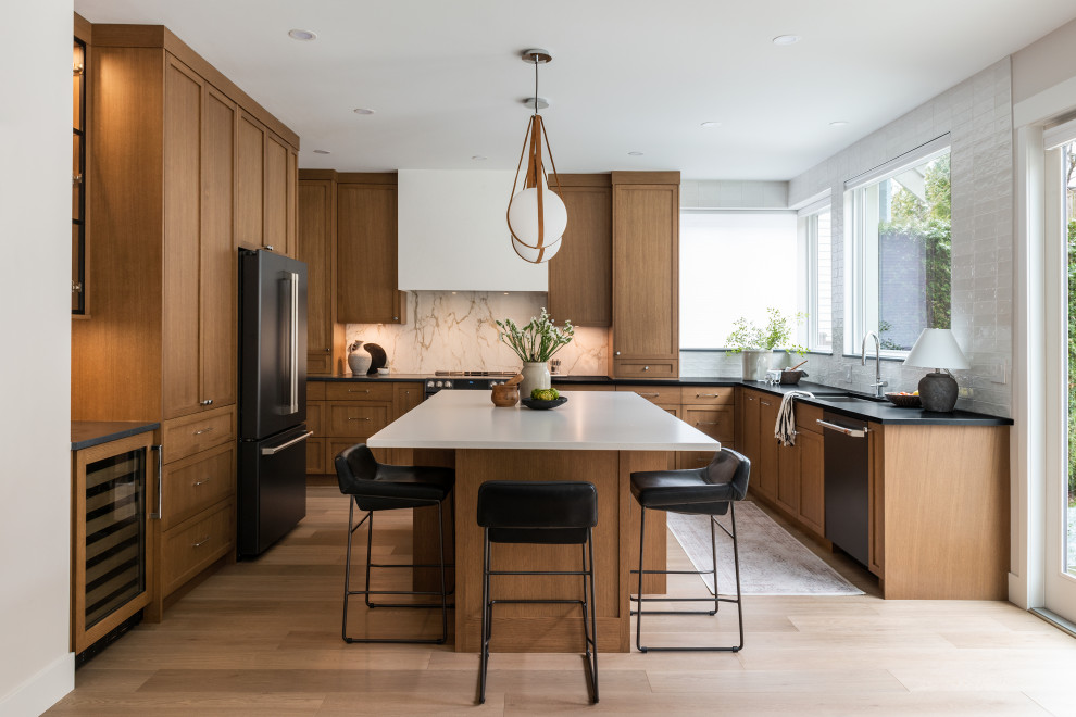 This is an example of a classic u-shaped kitchen in Vancouver with a submerged sink, shaker cabinets, medium wood cabinets, white splashback, stone slab splashback, black appliances, light hardwood flooring, an island, beige floors and black worktops.