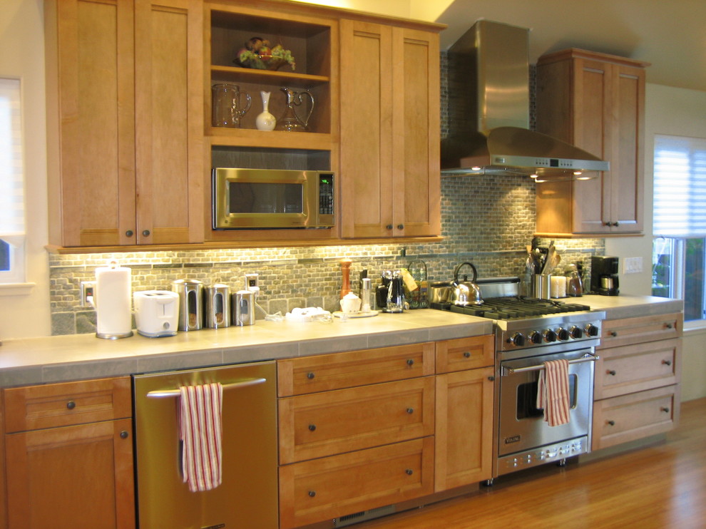 This is an example of a transitional l-shaped eat-in kitchen in San Francisco with shaker cabinets, light wood cabinets, tile benchtops, green splashback and stainless steel appliances.