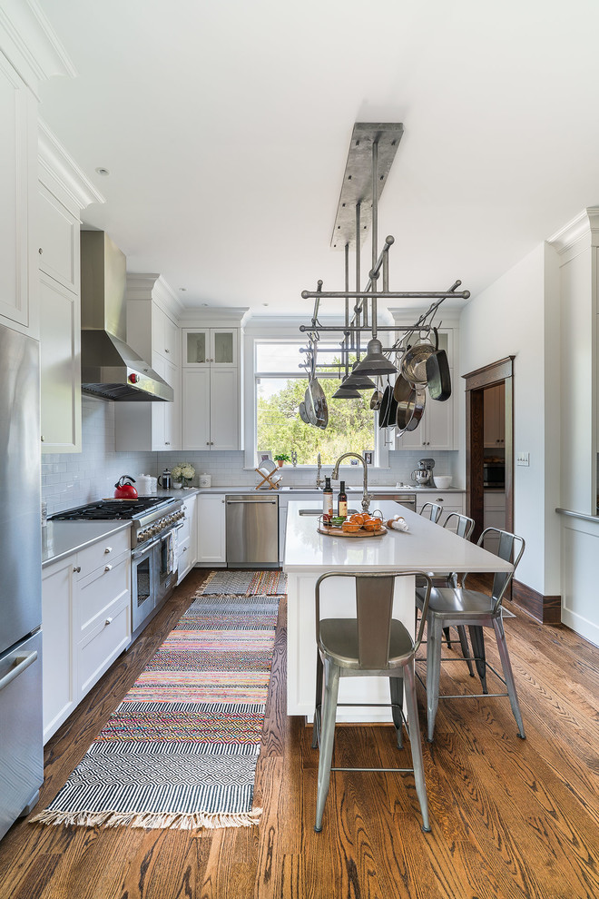 Inspiration for a mid-sized traditional kitchen in Austin with recessed-panel cabinets, white cabinets, subway tile splashback, stainless steel appliances, with island, brown floor, quartz benchtops, white splashback, medium hardwood floors and grey benchtop.