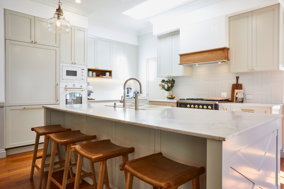 Large beach style l-shaped kitchen pantry in Sunshine Coast with an integrated sink, shaker cabinets, white cabinets, quartz benchtops, white splashback, subway tile splashback, black appliances, light hardwood floors, with island, brown floor and beige benchtop.