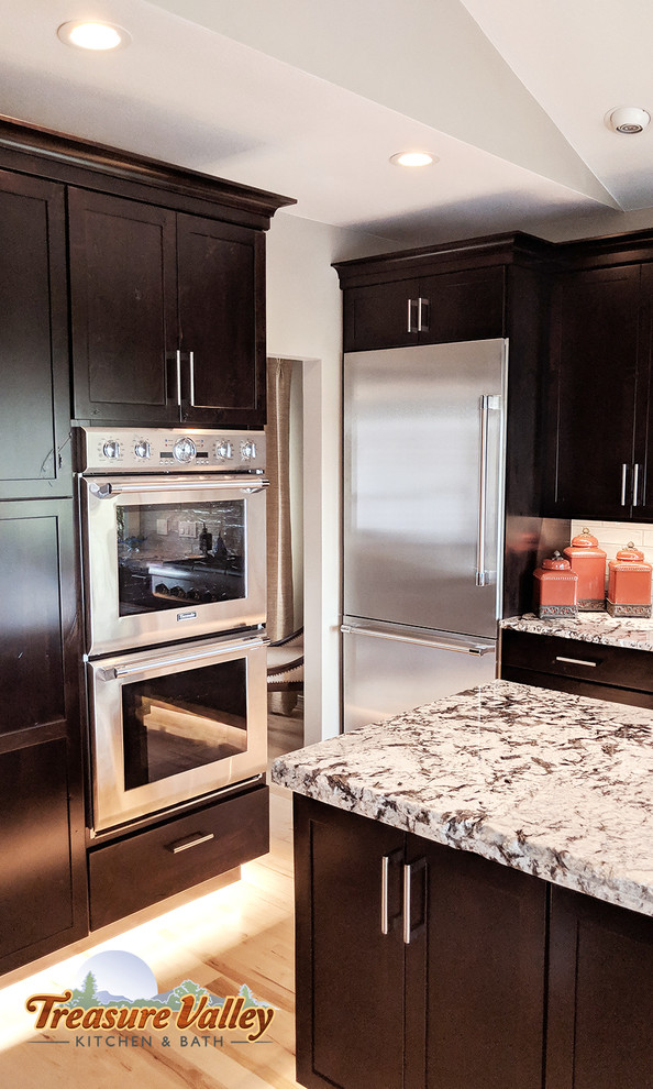Mid-sized transitional l-shaped light wood floor open concept kitchen photo in Boise with a farmhouse sink, shaker cabinets, granite countertops, glass tile backsplash, stainless steel appliances and an island