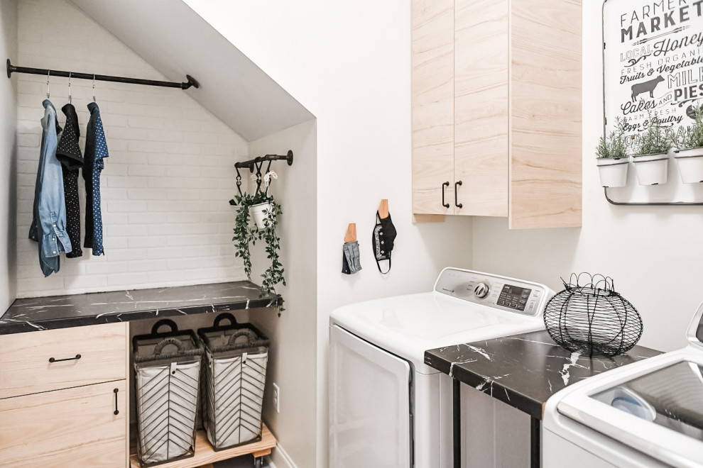 Mid-sized industrial l-shaped dedicated laundry room in Atlanta with flat-panel cabinets, light wood cabinets, laminate benchtops, white walls, linoleum floors, a side-by-side washer and dryer, multi-coloured floor, black benchtop and brick walls.