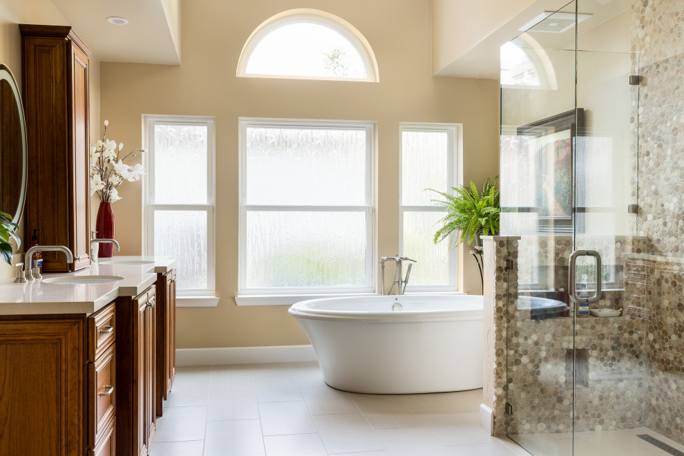 Large elegant master multicolored tile and pebble tile porcelain tile and beige floor freestanding bathtub photo with raised-panel cabinets, medium tone wood cabinets, a one-piece toilet, beige walls, an undermount sink, quartz countertops, a hinged shower door and white countertops