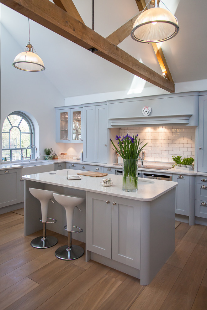 Design ideas for a mid-sized country l-shaped eat-in kitchen in Hertfordshire with quartz benchtops, with island, grey cabinets, white splashback, stainless steel appliances, medium hardwood floors, brown floor, grey benchtop, a farmhouse sink, shaker cabinets and subway tile splashback.