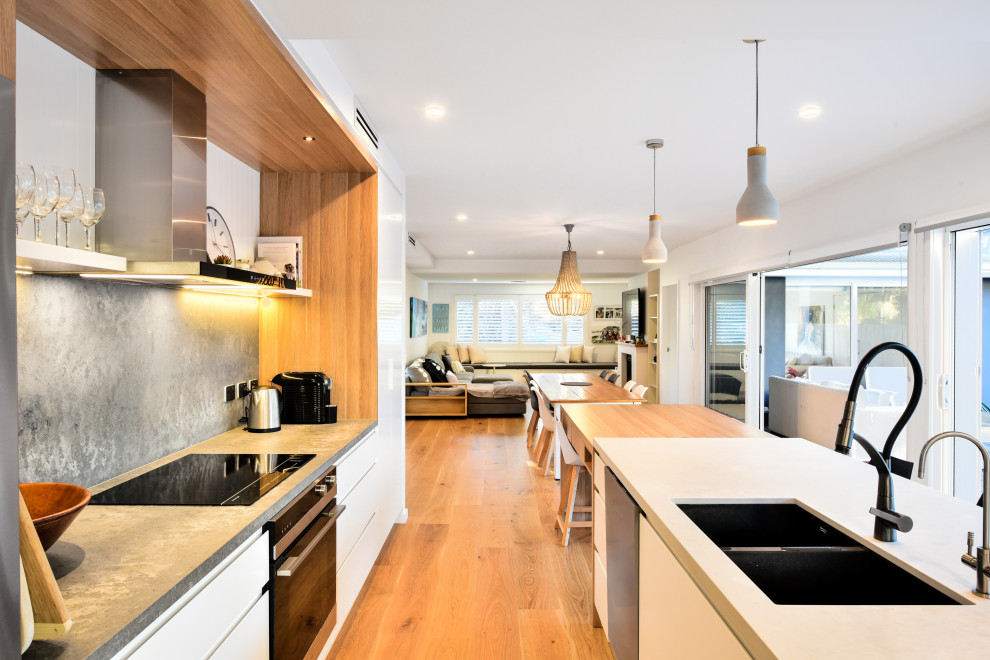 Photo of a large contemporary galley open plan kitchen in Sydney with a double-bowl sink, flat-panel cabinets, white cabinets, grey splashback, black appliances, a peninsula, brown floor and grey benchtop.