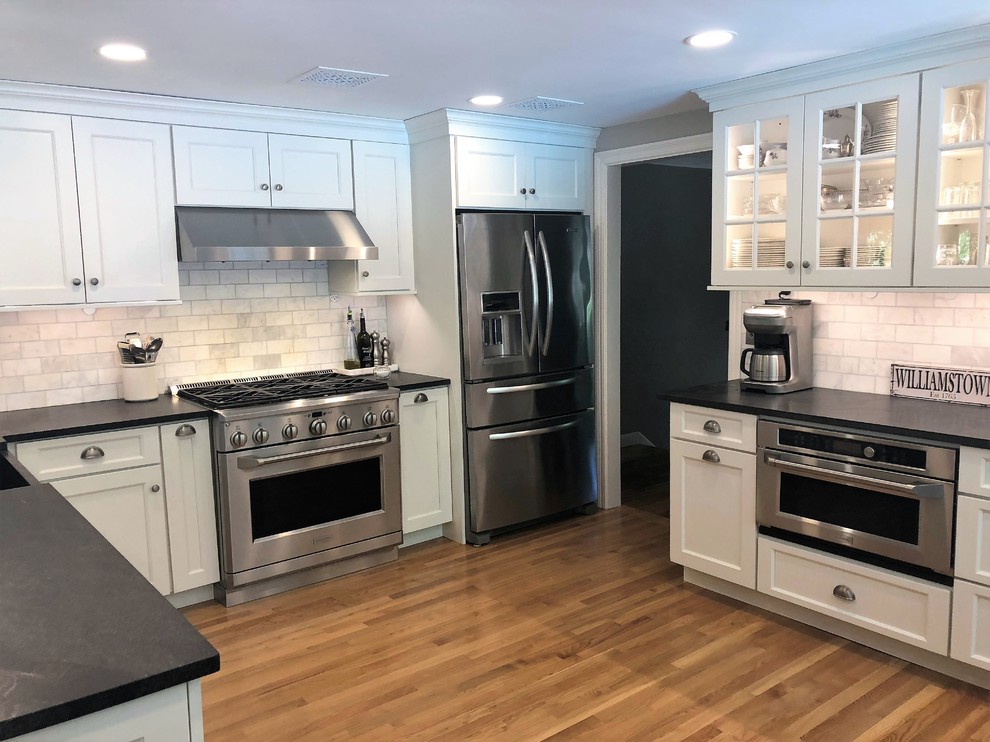 Mid-sized traditional l-shaped separate kitchen in Boston with a farmhouse sink, shaker cabinets, white cabinets, granite benchtops, white splashback, marble splashback, stainless steel appliances, medium hardwood floors, no island, beige floor and black benchtop.