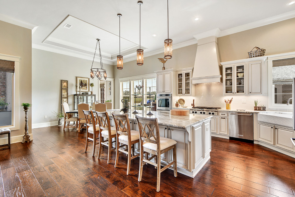 Design ideas for a large transitional l-shaped open plan kitchen in New Orleans with a farmhouse sink, beaded inset cabinets, white cabinets, marble benchtops, beige splashback, ceramic splashback, stainless steel appliances, medium hardwood floors, with island, brown floor and grey benchtop.