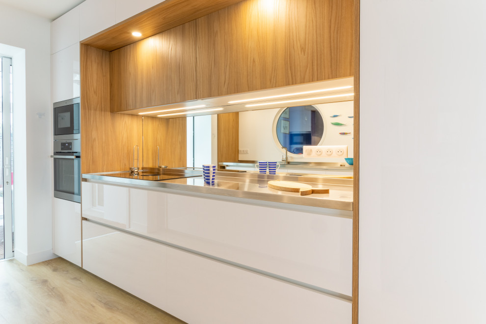 Exempel på ett litet maritimt kök, med en integrerad diskho, luckor med profilerade fronter, vita skåp, bänkskiva i rostfritt stål, spegel som stänkskydd, rostfria vitvaror och ljust trägolv