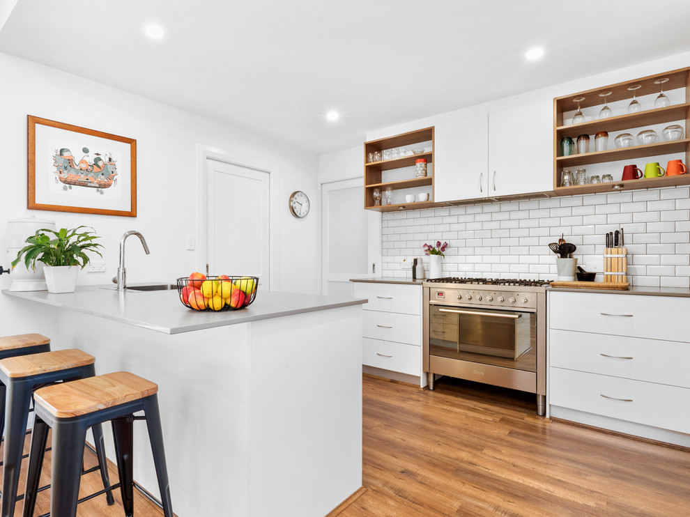 Small contemporary galley open plan kitchen in Perth with a single-bowl sink, white cabinets, solid surface benchtops, white splashback, subway tile splashback, stainless steel appliances, laminate floors, brown floor, flat-panel cabinets and a peninsula.