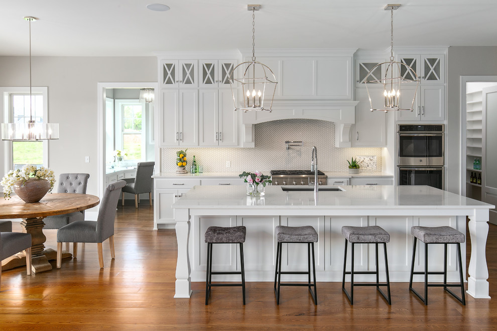 Inspiration for a transitional galley eat-in kitchen in Kansas City with an undermount sink, shaker cabinets, white cabinets, white splashback, mosaic tile splashback, stainless steel appliances, medium hardwood floors, with island, brown floor and white benchtop.