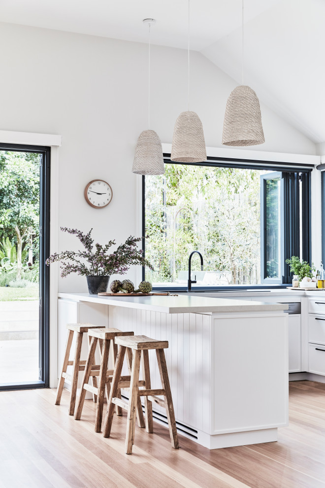 Photo of a medium sized coastal u-shaped kitchen/diner in Sydney with a double-bowl sink, shaker cabinets, white cabinets, engineered stone countertops, window splashback, stainless steel appliances, light hardwood flooring, a breakfast bar and grey worktops.