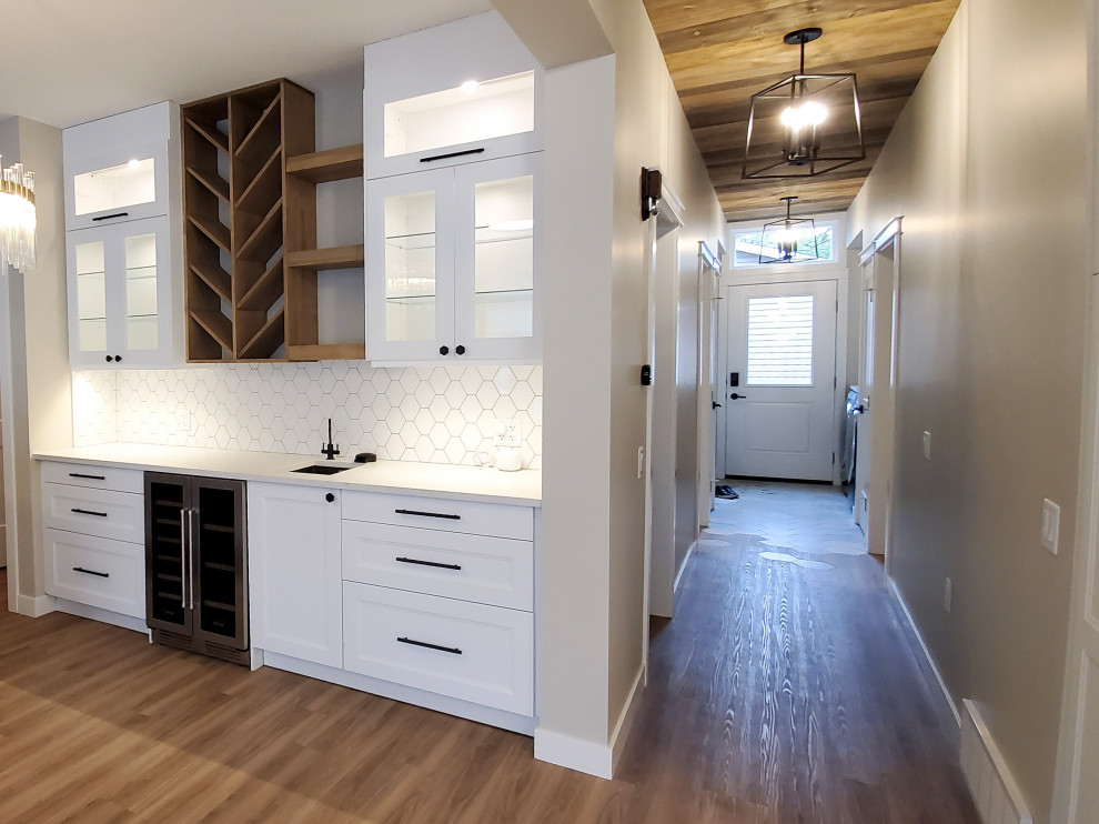 Photo of a mid-sized country single-wall wet bar in Edmonton with an undermount sink, beaded inset cabinets, white cabinets, quartz benchtops, white splashback, porcelain splashback, vinyl floors, brown floor and white benchtop.