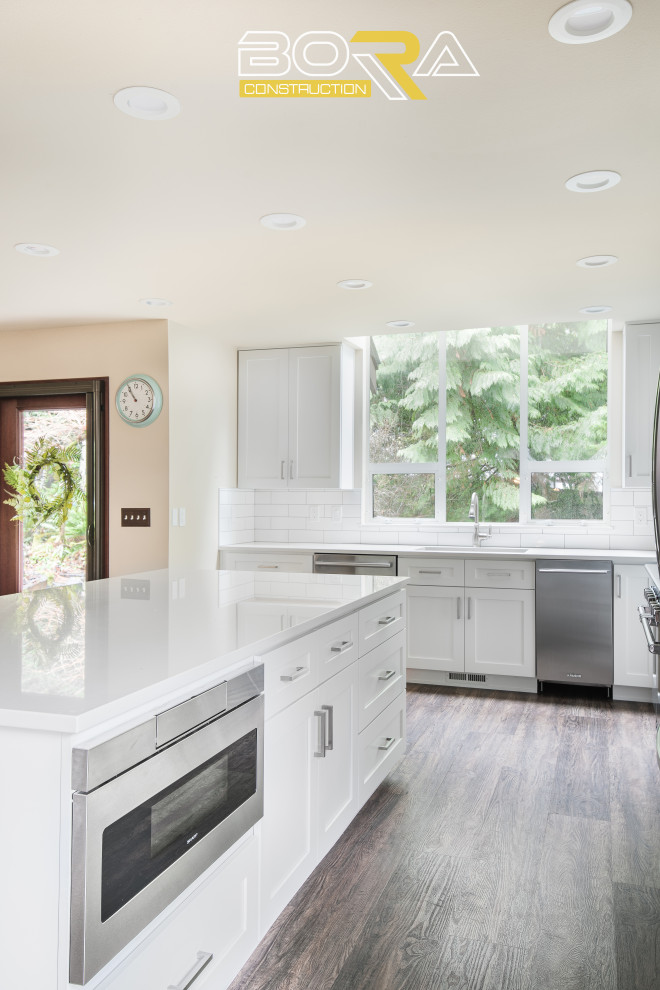 Inspiration for a large transitional l-shaped eat-in kitchen in Seattle with an undermount sink, shaker cabinets, white cabinets, quartz benchtops, white splashback, ceramic splashback, stainless steel appliances, medium hardwood floors, with island, brown floor and white benchtop.