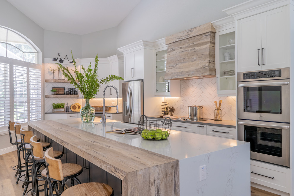 This is an example of a large country galley open plan kitchen in Miami with a farmhouse sink, shaker cabinets, white cabinets, quartzite benchtops, white splashback, subway tile splashback, stainless steel appliances, vinyl floors, with island, beige floor and grey benchtop.