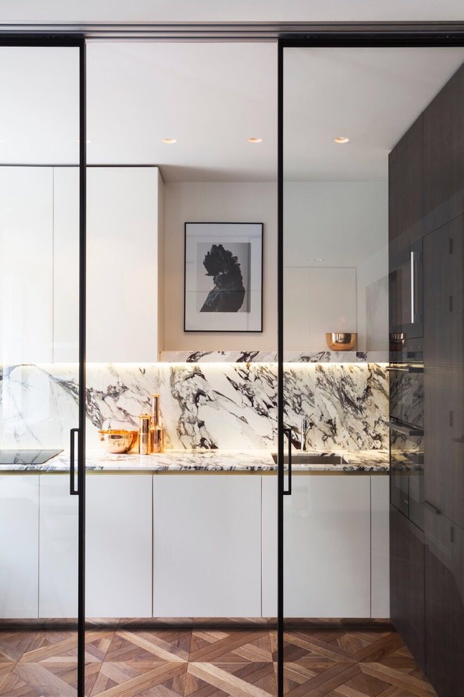 Photo of a mid-sized traditional single-wall open plan kitchen in London with a drop-in sink, flat-panel cabinets, white cabinets, marble benchtops, white splashback, marble splashback, white appliances, dark hardwood floors, no island and brown floor.