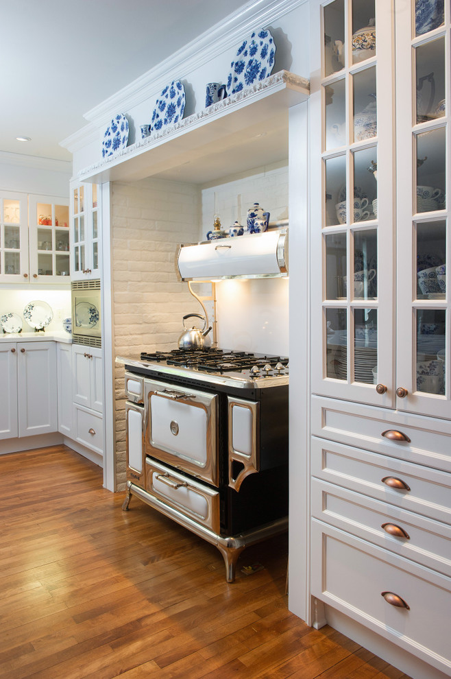 Photo of a large traditional u-shaped eat-in kitchen in Montreal with a drop-in sink, glass-front cabinets, white splashback, ceramic splashback, white appliances, medium hardwood floors, with island and white cabinets.