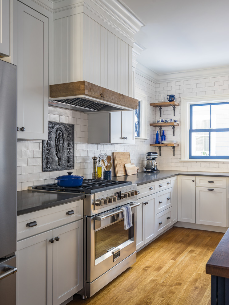 Photo of a mid-sized country l-shaped eat-in kitchen in Boston with a farmhouse sink, shaker cabinets, white cabinets, quartz benchtops, white splashback, subway tile splashback, stainless steel appliances, medium hardwood floors and with island.