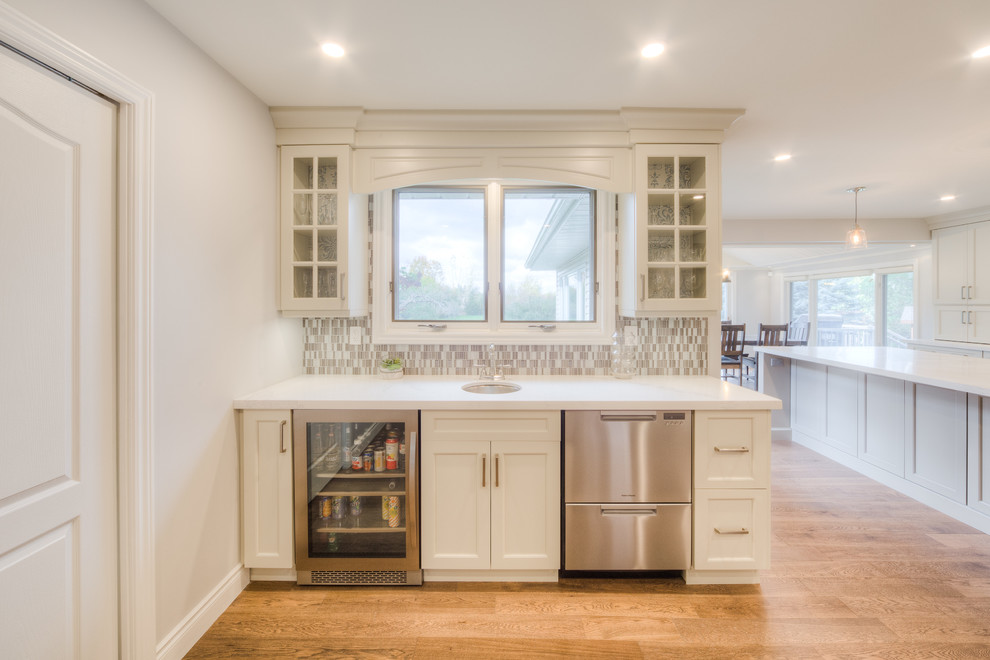 Inspiration for a mid-sized transitional galley light wood floor and beige floor open concept kitchen remodel in Toronto with a double-bowl sink, recessed-panel cabinets, white cabinets, solid surface countertops, white backsplash, porcelain backsplash, stainless steel appliances, an island and white countertops