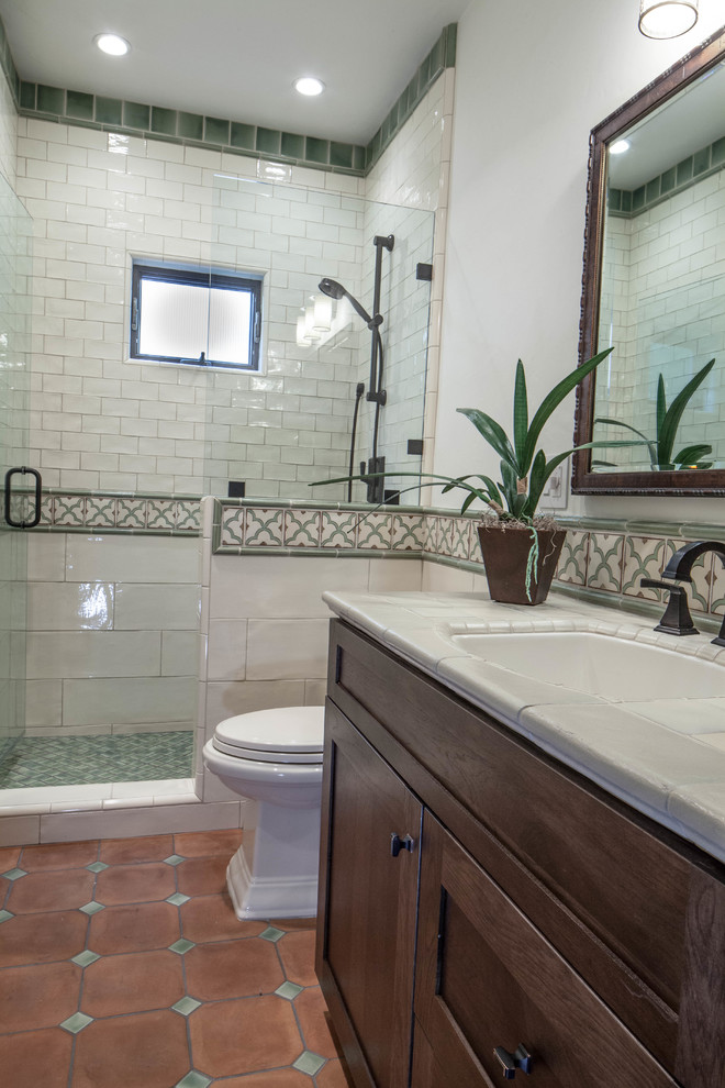 This is an example of a mid-sized eclectic kids bathroom in San Luis Obispo with shaker cabinets, medium wood cabinets, a one-piece toilet, beige tile, ceramic tile, beige walls, terra-cotta floors, an undermount sink, tile benchtops, red floor, a hinged shower door and beige benchtops.