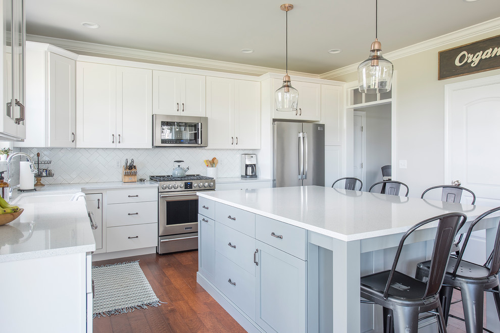 Example of a mid-sized transitional l-shaped medium tone wood floor and brown floor open concept kitchen design with a farmhouse sink, shaker cabinets, white cabinets, quartzite countertops, white backsplash, subway tile backsplash, stainless steel appliances and an island