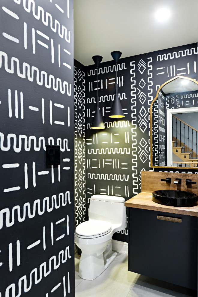 Inspiration for a mid-sized eclectic powder room in Toronto with flat-panel cabinets, black cabinets, a one-piece toilet, black walls, porcelain floors, a vessel sink, wood benchtops, white floor, brown benchtops, a built-in vanity and wallpaper.
