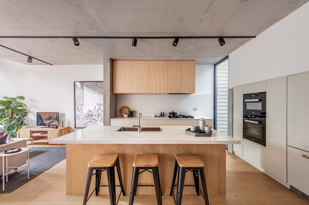 This is an example of a contemporary l-shaped kitchen in Sydney with an undermount sink, flat-panel cabinets, beige cabinets, black appliances, medium hardwood floors, with island, brown floor and white benchtop.