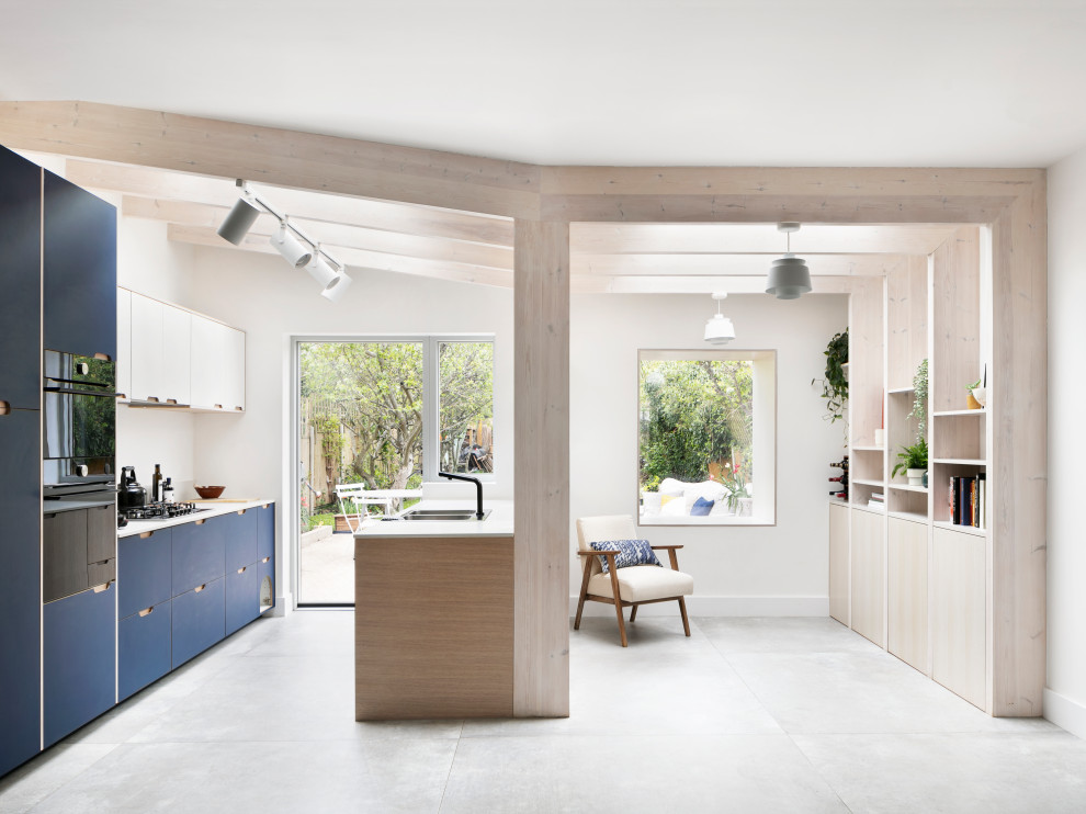 This is an example of a mid-sized scandinavian galley open plan kitchen in London with a drop-in sink, flat-panel cabinets, blue cabinets, solid surface benchtops, white splashback, granite splashback, black appliances, porcelain floors, no island, grey floor, white benchtop and exposed beam.