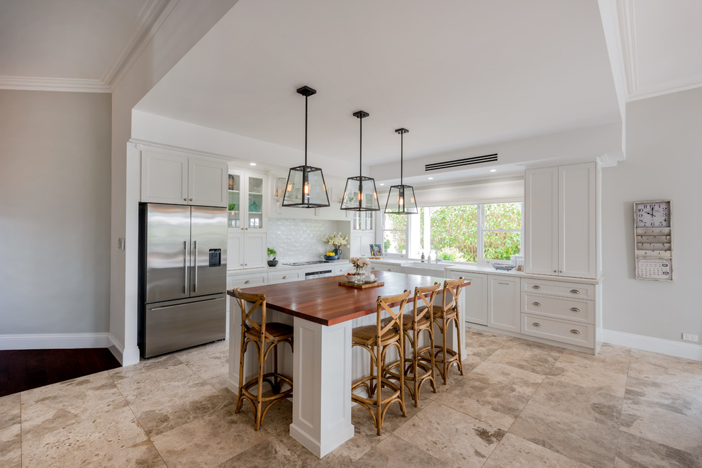 Inspiration for a large traditional l-shaped eat-in kitchen in Other with a farmhouse sink, shaker cabinets, white cabinets, wood benchtops, white splashback, cement tile splashback, black appliances, porcelain floors, with island, beige floor and white benchtop.