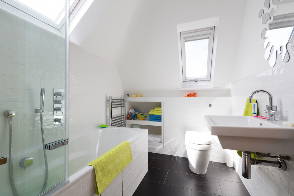 Inspiration for a contemporary kids bathroom in London with a wall-mount sink, open cabinets, white cabinets, a drop-in tub, a shower/bathtub combo, a wall-mount toilet, white tile, white walls and black floor.