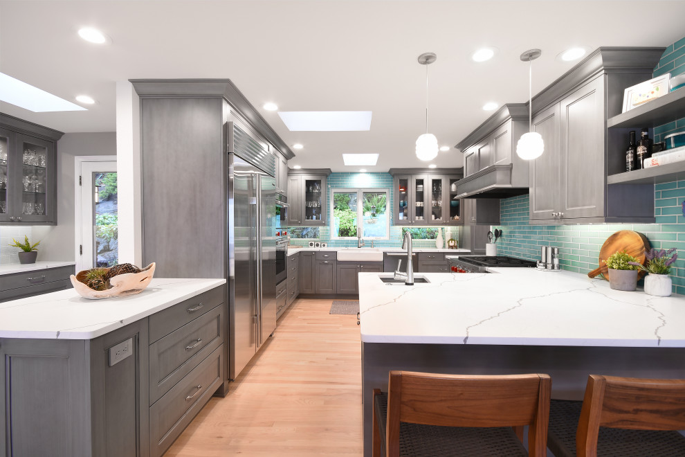 This is an example of a large transitional eat-in kitchen in Seattle with a farmhouse sink, recessed-panel cabinets, grey cabinets, quartz benchtops, blue splashback, ceramic splashback, stainless steel appliances, light hardwood floors, a peninsula, yellow floor and white benchtop.