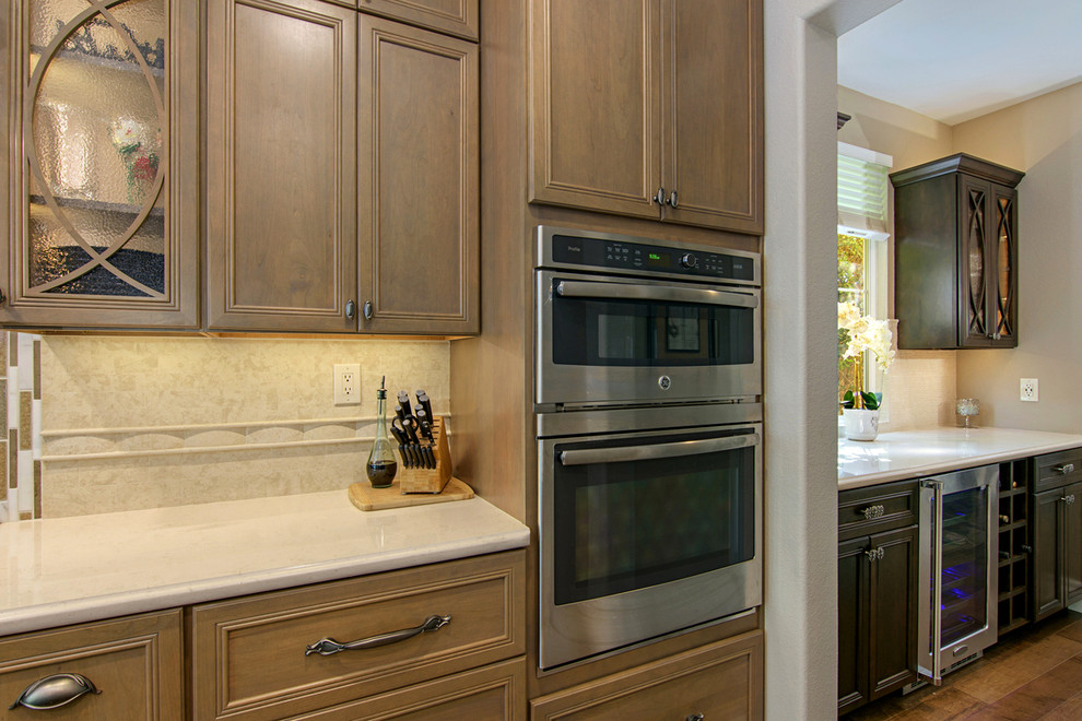 Example of a large transitional l-shaped medium tone wood floor and brown floor open concept kitchen design in San Diego with a farmhouse sink, recessed-panel cabinets, medium tone wood cabinets, beige backsplash, paneled appliances, an island, quartz countertops and travertine backsplash