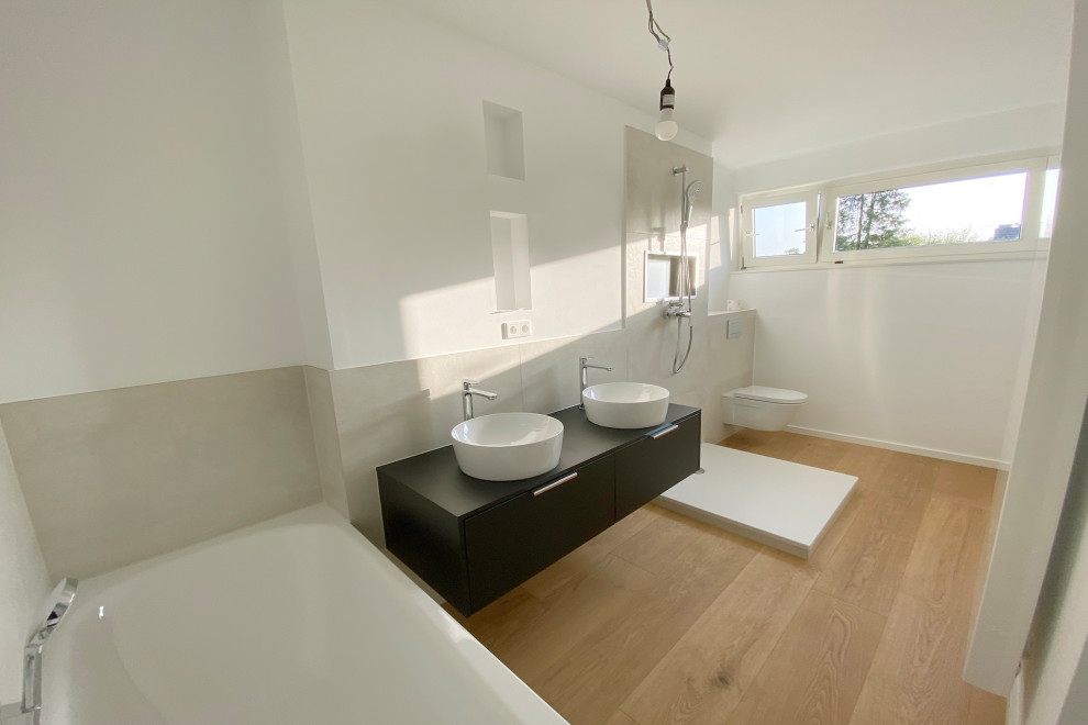 Badezimmer With flächenbündigen Schrankfronten, dunklen Holzschränken, Einbaubadewanne, Duschbadewanne, Wandtoilette mit Spülkasten, beigen Fliesen, Keramikfliesen, hellem Holzboden, Waschtisch aus Holz, schwarzer Waschtischplatte, Doppelwaschbecken and eingebautem Waschtisch in Dusseldorf