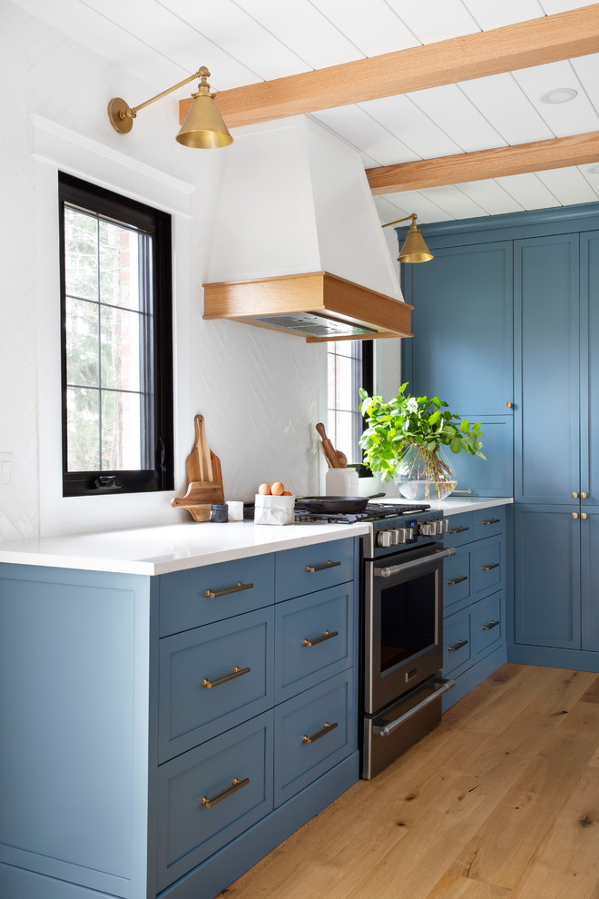 Design ideas for a mid-sized country l-shaped eat-in kitchen in Montreal with a farmhouse sink, shaker cabinets, blue cabinets, quartz benchtops, white splashback, porcelain splashback, stainless steel appliances, light hardwood floors, with island, brown floor and white benchtop.