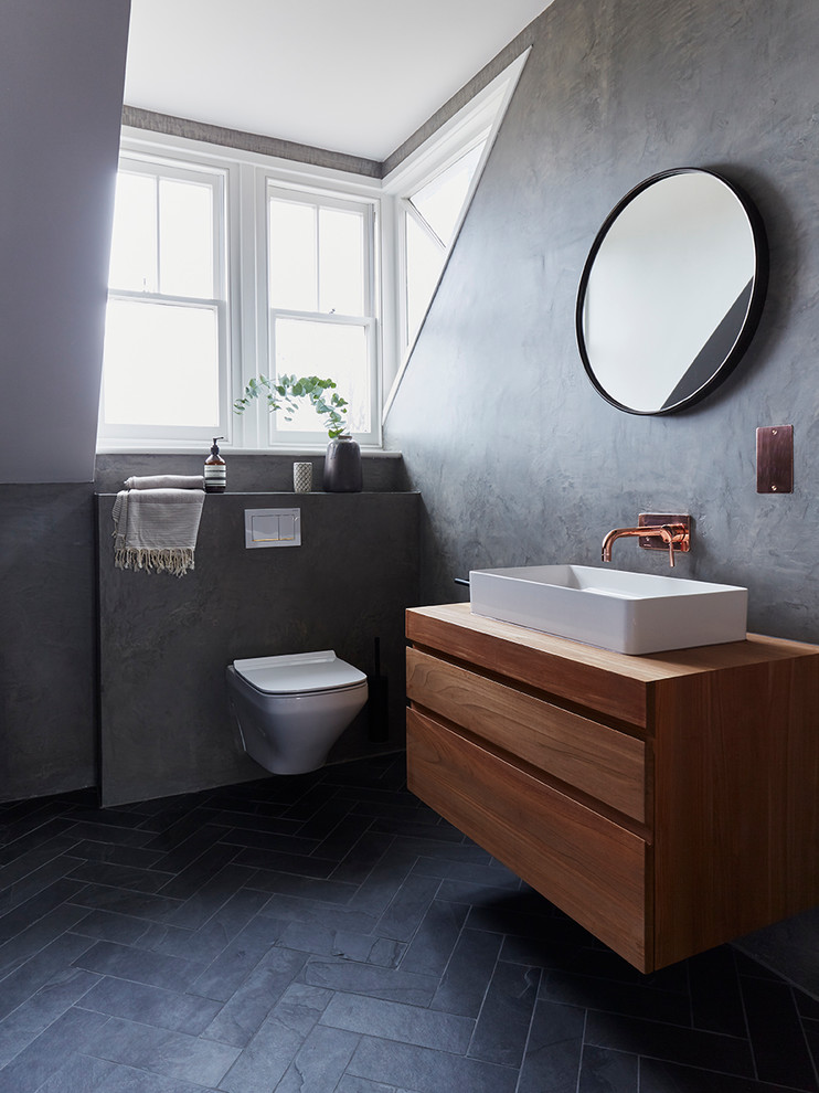 This is an example of a modern 3/4 bathroom in Hertfordshire with flat-panel cabinets, medium wood cabinets, a wall-mount toilet, grey walls, a vessel sink, wood benchtops, black floor and brown benchtops.