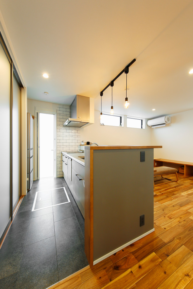 This is an example of a mid-sized industrial single-wall open plan kitchen in Tokyo Suburbs with a peninsula, an integrated sink, flat-panel cabinets, grey cabinets, stainless steel benchtops, white splashback, ceramic splashback, panelled appliances, terrazzo floors, grey floor and brown benchtop.