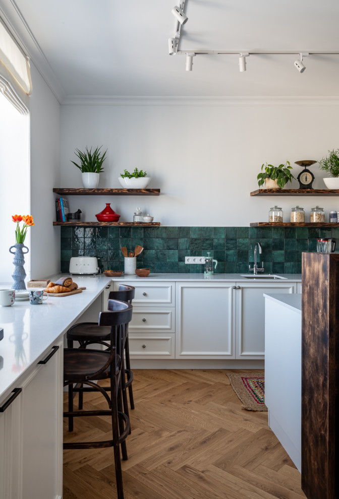 Foto på ett vintage vit l-kök, med en undermonterad diskho, luckor med infälld panel, vita skåp, grönt stänkskydd, mellanmörkt trägolv, en köksö och brunt golv