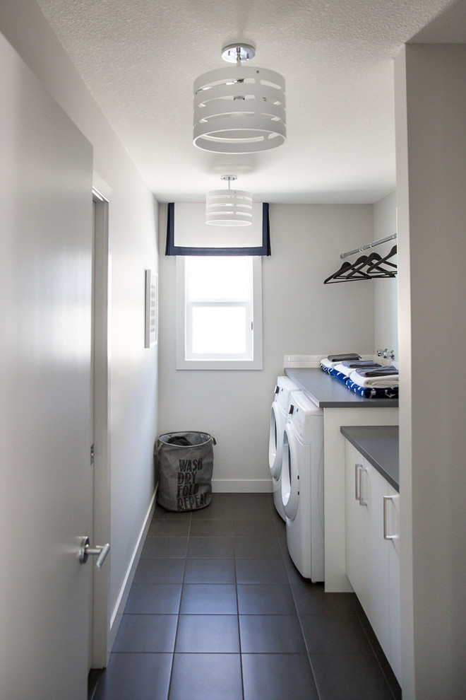 This is an example of a mid-sized contemporary galley dedicated laundry room in Calgary with flat-panel cabinets, white cabinets, quartz benchtops, white walls, ceramic floors, a side-by-side washer and dryer and grey floor.