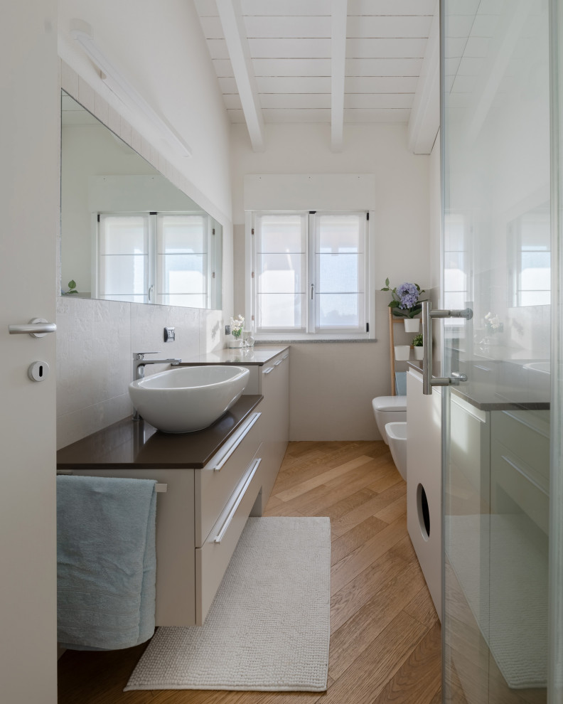 На фото: узкая и длинная ванная комната среднего размера в скандинавском стиле с плоскими фасадами, бежевыми фасадами, угловым душем, раздельным унитазом, белой плиткой, керамической плиткой, белыми стенами, светлым паркетным полом, душевой кабиной, настольной раковиной, столешницей из искусственного кварца, бежевым полом, душем с раздвижными дверями, бежевой столешницей, тумбой под одну раковину, встроенной тумбой и балками на потолке с