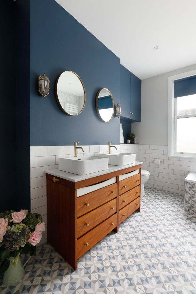 This is an example of a large contemporary master bathroom in Sussex with furniture-like cabinets, medium wood cabinets, a corner tub, an open shower, a one-piece toilet, white tile, ceramic tile, blue walls, porcelain floors, a vessel sink, engineered quartz benchtops, blue floor, an open shower, white benchtops, a double vanity and a freestanding vanity.