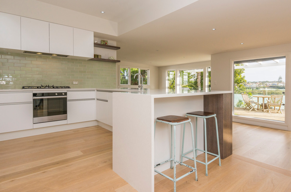 Mid-sized contemporary u-shaped kitchen in Auckland with an undermount sink, flat-panel cabinets, white cabinets, quartz benchtops, green splashback, subway tile splashback, stainless steel appliances and light hardwood floors.