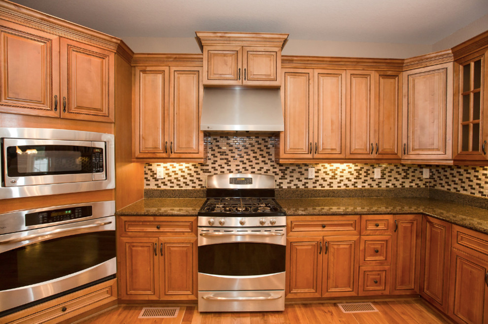 Medium tone wood floor and brown floor kitchen photo with a double-bowl sink, glass-front cabinets, medium tone wood cabinets, granite countertops, black backsplash, ceramic backsplash, stainless steel appliances, an island and brown countertops