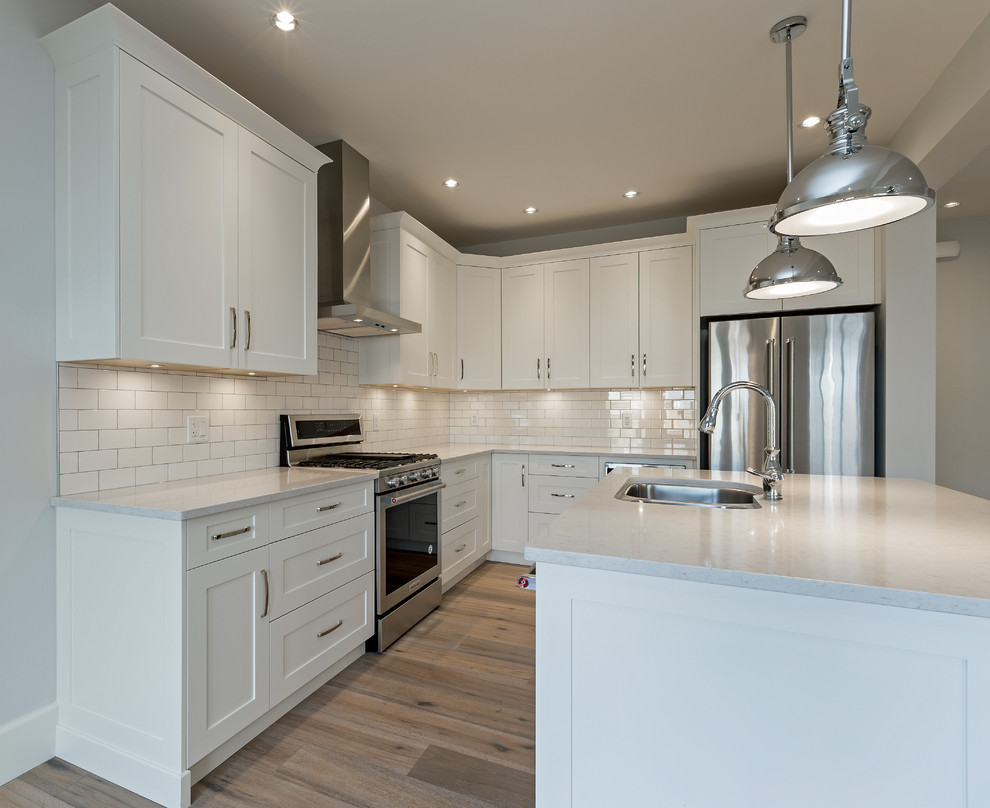 Mid-sized transitional l-shaped light wood floor open concept kitchen photo with an undermount sink, shaker cabinets, white cabinets, quartz countertops, white backsplash, subway tile backsplash, stainless steel appliances, an island and white countertops