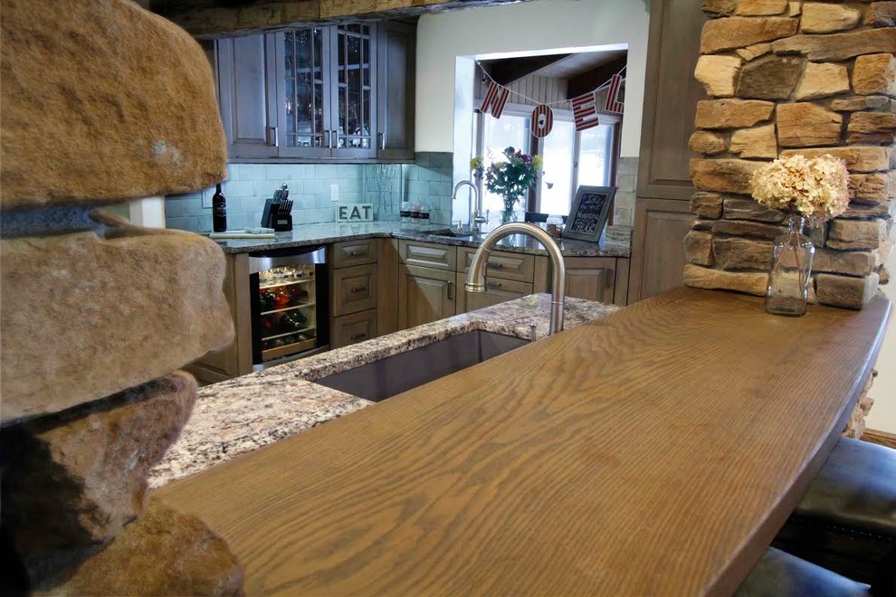 Example of a large mountain style u-shaped medium tone wood floor and brown floor open concept kitchen design with a single-bowl sink, raised-panel cabinets, gray cabinets, granite countertops, gray backsplash, brick backsplash, stainless steel appliances and an island